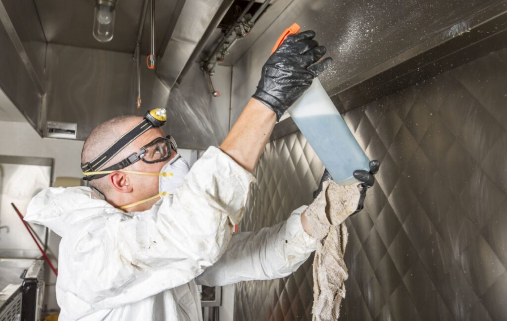 A commercial cleaner wearing a mask sprays a degreasing solution onto the range hood in an industrial kitchen to clean it.