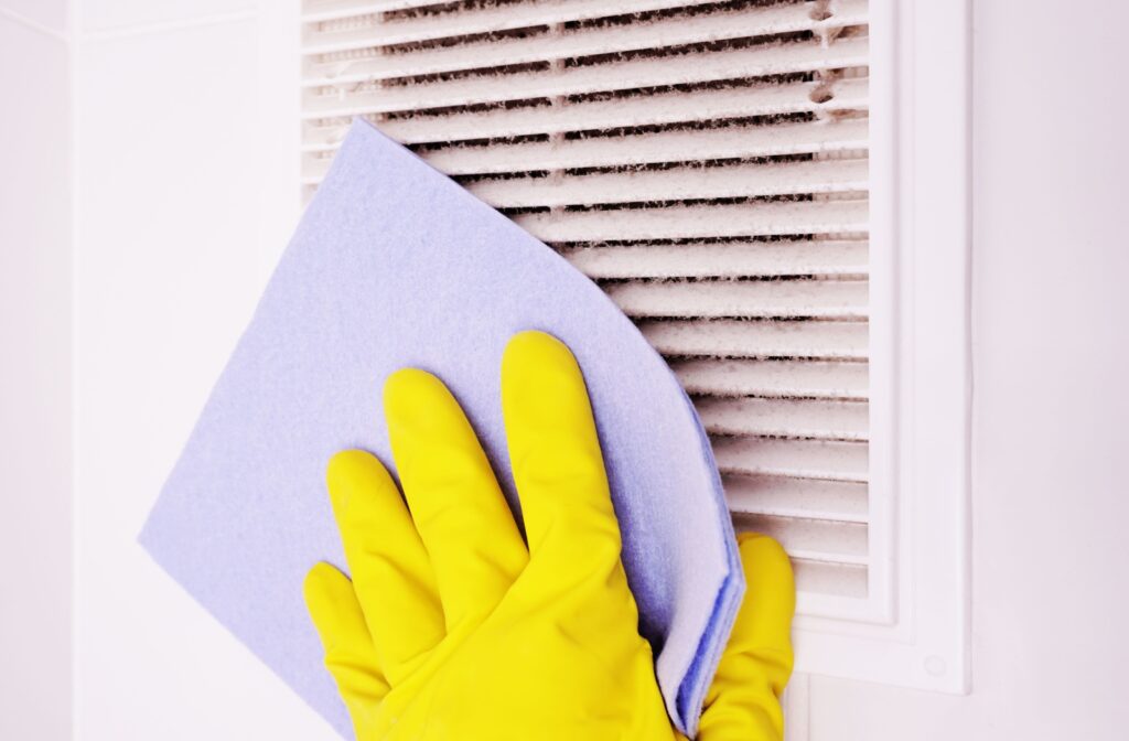 A hand in a yellow glove cleaning a dusty vent with a lavender cloth.