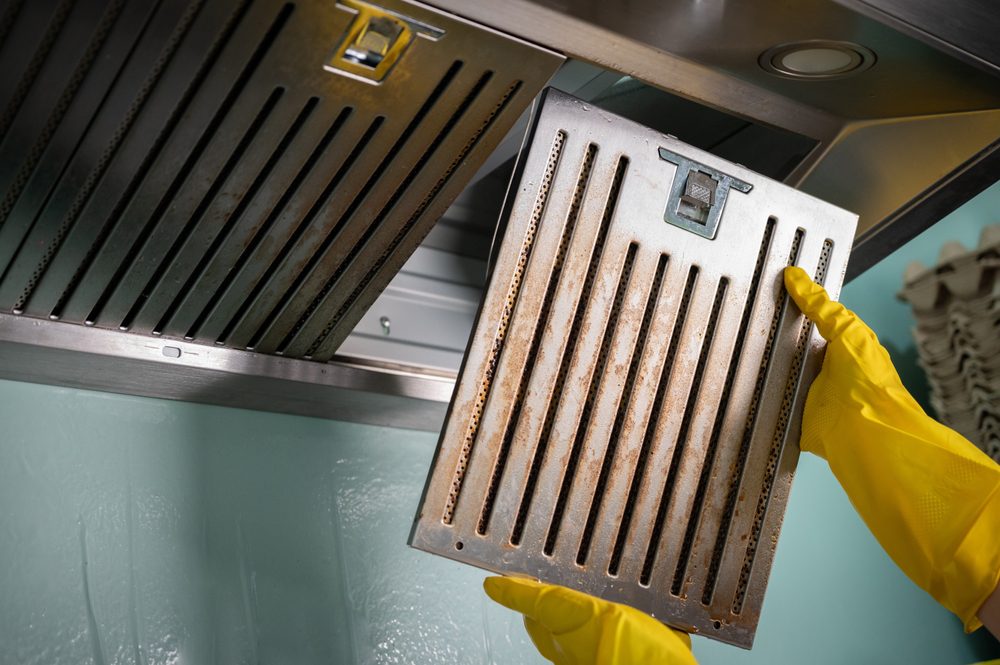 Hands wearing yellow rubber gloves remove a greasy metal filter from a stainless steel kitchen range hood during cleaning.