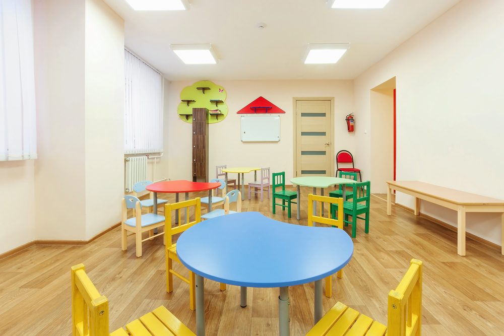 Bright preschool classroom with small colorful tables and chairs arranged on a wooden floor, soft lighting, and child-friendly wall decorations.