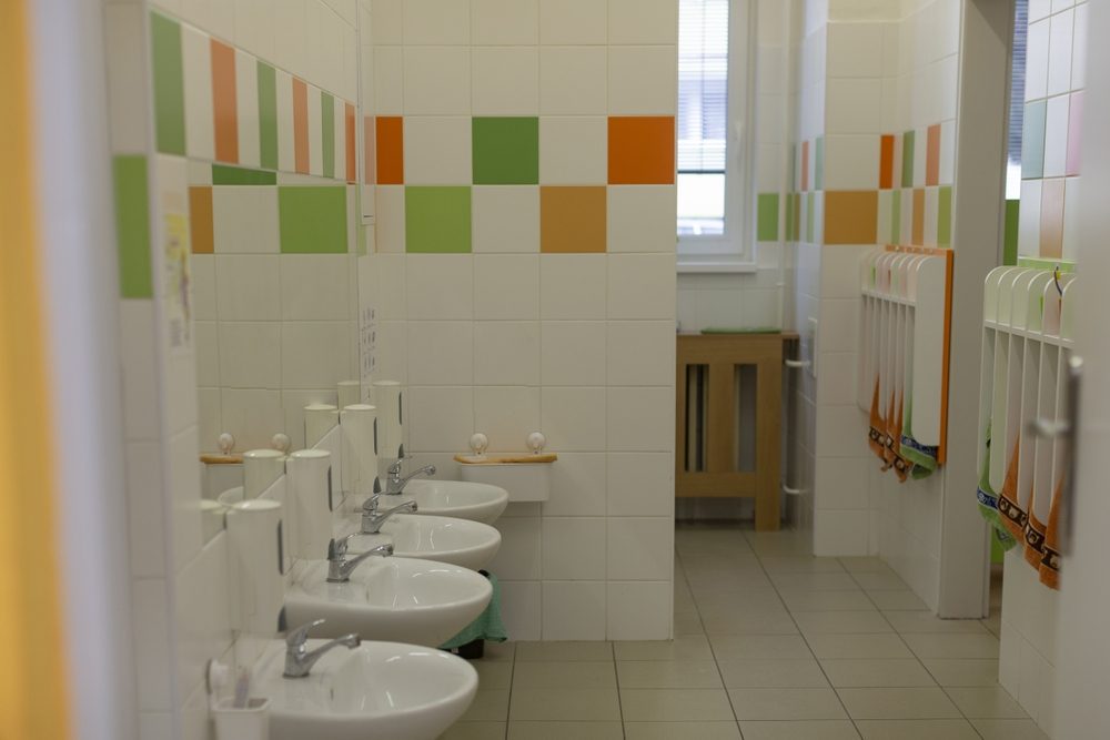 Clean children’s bathroom with multiple low sinks, white tiled walls with green and orange accents, and small towels hanging on racks.